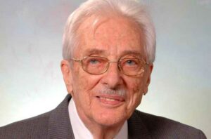 Man with white hair and glasses in dark grey suit.